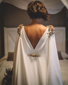 the back of a woman's white dress with gold flowers on it, standing in front of a bed