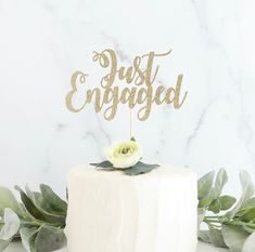 a white cake topped with a gold just engaged cake topper next to greenery