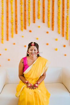 a woman in a yellow sari sitting on a white couch with flowers hanging from the wall behind her