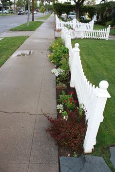 a white picket fence next to a sidewalk with flowers growing on the curb and grass around it