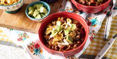 two red bowls filled with chili and avocado on top of a wooden cutting board