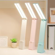 three different colored lights on top of a white desk next to a plant and bookshelf