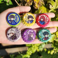 four different colored bottle caps in the palm of someone's hand, with anime characters on them