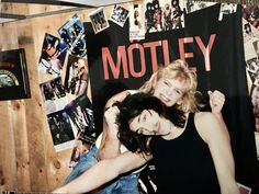 two women standing next to each other in front of a wooden wall covered with photos