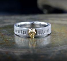 This gold ring is a narrow, elegant band that combines symbolism and style. Its minimalist design belies its deep meaning, capturing attention with an intriguing central motif: a finely sculpted skull that sits precisely on the surface of the ring. The skull, rendered in detail, evokes a sense of mystery and a constant reminder of mortality. On the outside of the band, the phrase "Memento Mori" is engraved in careful, well-defined letters, reminding the wearer of the impermanence of life. This inscription, meaning "Remember that you will die," serves as a philosophical reflection and a call to live with purpose and awareness. The shine of the gold adds a touch of luxury, while the simple design of the band ensures that the ring is comfortable to wear. This ring is ideal both as a meaningfu Symbolic White Gold Skull Ring For Anniversary, Symbolic Hallmarked Skull Ring For Anniversary, Symbolic Stainless Steel Engraved Ring For Anniversary, Symbolic Engraved White Gold Skull Ring, Gift Engraved Stainless Steel Skull Ring, Gold Engraved Skull Ring For Anniversary, Live With Purpose, Deep Meaning, Memento Mori