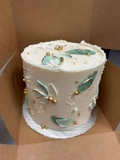 a white cake sitting in a box on top of a cardboard surface with gold beads and leaves