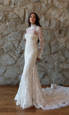 a woman standing in front of a stone wall wearing a wedding dress with long sleeves