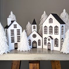 paper houses are lined up on a table with trees in the foreground and behind them is a gray wall