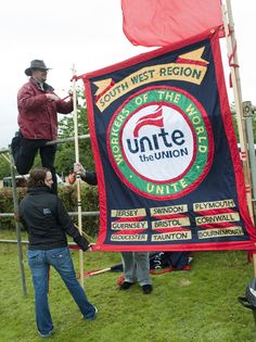 two people standing on top of a sign in the grass with one person holding a flag