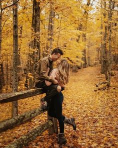 a man and woman kissing on a wooden fence in the woods with leaves all around them