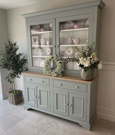 a gray china cabinet with flowers and plates on the glass doors, next to a potted plant