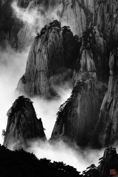 black and white photograph of mountains covered in fog with trees on the top, surrounded by low hanging clouds