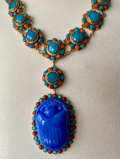 A vintage striking and beautiful  1930's Egyptian Revival coral and turquoise glass Scarab drop necklace. A single row of turquoise and coral glass cabochon rosettes, 15 in total, with a pendant of a further rosette and a large turquoise glass Scarab Beetle with a surround of turquoise and coral glass cabochons. The metal mounts are gilt metal and the cabochons collet set. A lovely clasp set with 3 glass turquoise cabochons, hook and ring clasp secures. Length of 17 inch with 4.1/2 inch drop. In Traditional Blue Cabochon Necklace, Traditional Blue Cabochon Necklaces, Antique Blue Cabochon Necklace, Scarab Amulet, Ancient Egyptian Jewelry Necklaces, Art Nouveau Jewelry Egyptian, Scarab Beetle Egyptian Jewelry, Egyptian Scarab Necklace, Czech Jewelry