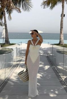 a woman in a white dress and hat walking on a boardwalk near the ocean with palm trees