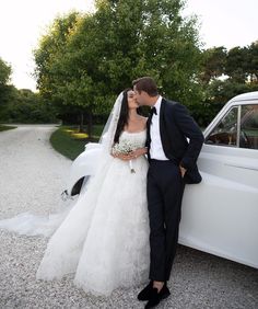 a bride and groom kissing in front of a white car on gravel road next to trees