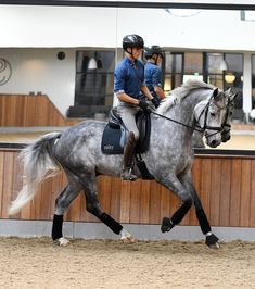 two people riding on the back of a gray horse in an indoor arena with wooden walls