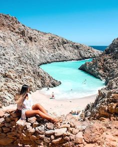 a woman is sitting on rocks near the beach and looking out at the blue water