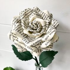 a white rose made out of book pages sitting in a vase with green leaves on the side