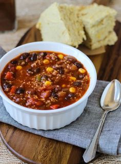 a white bowl filled with chili and corn on top of a wooden cutting board next to bread