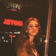 a woman wearing sunglasses standing in front of a store window at night with the lights on