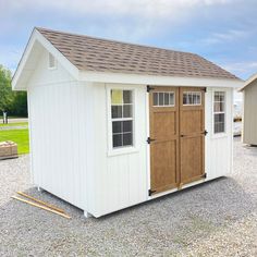 a small white shed with brown doors and shingles on the roof is sitting in gravel