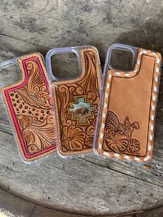 three cell phone cases sitting next to each other on a wooden bench, one has a leather case and the other has an embroidered design