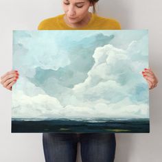 a woman holding up a painting with clouds in the sky