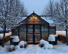 a small glass house in the middle of snow covered trees and bushes with lights on