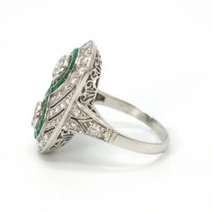 This jaw dropping ring, an Art Deco piece of art likely made in the 1920s, is made of platinum and features two central bezel set diamonds ensconced by an infinity-style figure eight channel of square cut emeralds. The rest of the ring's top contains multiple angular swirls of diamonds that are separated by pierced cut-outs, giving a palpable sense of motion to the piece. The shoulders are beautifully pierced and the undercarriage—or gallery—is ornately filigreed. Metal: Platinum Main Stones: 2 Round Brilliant Diamonds (approx. 0.66 carat total, G color, SI1-SI2 clarity, natural, 4.4 mm) Accent Stones: 42 Round Brilliant Diamonds (approx. 0.82 carat total, F-H color, VS1-SI2 clarity, natural, 1.0 - 2.3 mm) & 40 Square Cut Emeralds (approx. 0.40 carat total, natural) Ring Size: 7.25 (can be Art Deco Diamond White Jewelry With Bezel Setting, Art Deco Diamond Jewelry With Bezel Setting, Diamond White Art Deco Jewelry With Bezel Setting, Art Deco White Gold Diamond Ring With Bezel Setting, Art Deco Ring With Bezel Setting, Art Deco Silver Diamond Ring With Bezel Setting, Silver Art Deco Diamond Ring With Bezel Setting, Art Deco Platinum Jewelry With Bezel Setting, Platinum Art Deco Jewelry With Bezel Setting