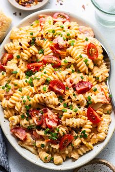 a bowl full of pasta salad with tomatoes and green onions on the side next to a glass of water