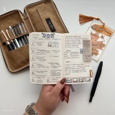 a hand holding an open notebook with writing on it next to other items including pens and pencils