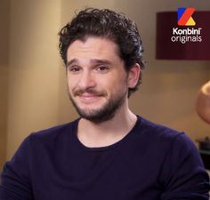 a man with curly hair and beard sitting in front of a tv screen wearing a blue shirt