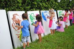 several children are drawing on canvases in front of a fence with trees and bushes behind them