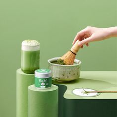 a person holding a wooden brush over a green container on top of a table next to other items