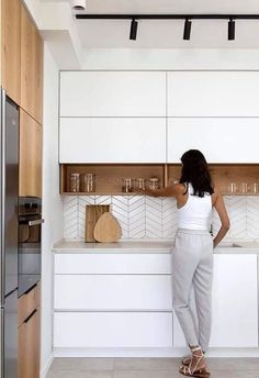 a woman standing at the counter in a kitchen with white cupboards and drawers on both sides