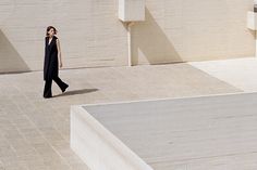 a woman is walking in the middle of an empty courtyard with white walls and concrete steps