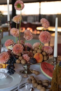 an arrangement of fruit and flowers on a table with candles in the background, including watermelon