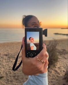 a woman holding up a camera to take a photo on the beach with her cell phone