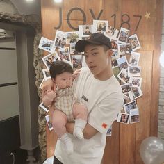 a man holding a baby in front of a wooden wall with photos on it and balloons