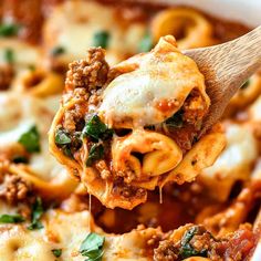 a wooden spoon full of pasta with meat and spinach on it, being lifted from a casserole dish