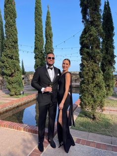 a man and woman standing next to each other in front of some trees with lights strung over them