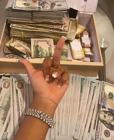 a person's hand pointing at stacks of money in a box on top of a table