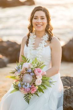 a woman in a wedding dress is holding a bouquet on the rocks by the water