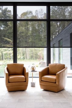 two brown leather chairs sitting in front of large windows with trees out the window behind them