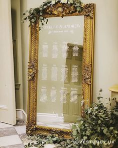 an ornate gold framed wedding seating chart with greenery on the floor and in front of it