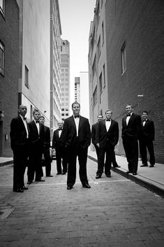 a group of men in tuxedos are standing on the sidewalk near tall buildings