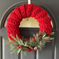 a red knitted wreath hanging on the front door with pine cones and evergreens