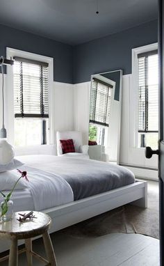 a white bed sitting next to two windows with shutters on each side and a table in front of it