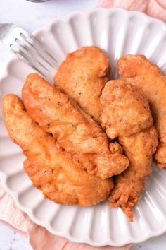 some fried chicken on a white plate with a fork and sauce in the bowl next to it