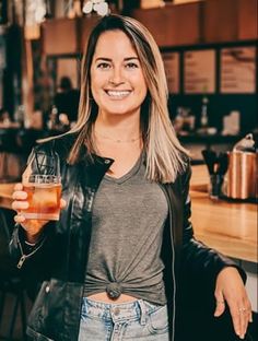 a woman holding a drink in her right hand and smiling at the camera while standing next to a bar